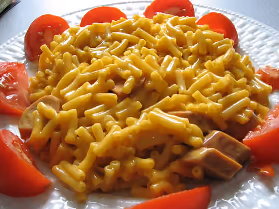 Makaroni saus keju — Kraft Dinner (macaroni and cheese) with added cheddar and sliced vegetarian hot dogs arranged on a plate and bordered by tomato wedges.