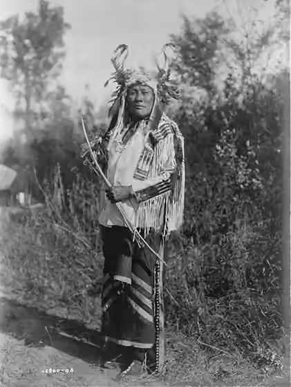 Hidatsa — Long Time Dog, full-length portrait, standing, facing front, wearing headress, buckskin shirt, and leggings with brass bells.