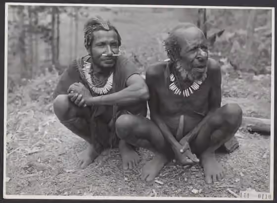 Suku Ngalum — Collectie / Archief: Kantoor voor Voorlichting en Radio Omroep Nederlands Nieuw Guinea Reportage / Serie: Expeditie Sterrengebergte en Julianatop (1959) - Nieuw-Guinea Beschrijving: [twee Papoeamannen zitten gehurkt naast elkaar] Datum:…